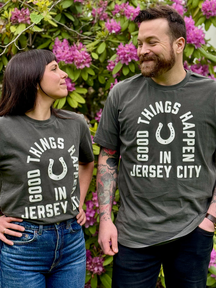 Man and woman wearing Good Things Happen in Jersey City shirt standing outdoors with purple flowers