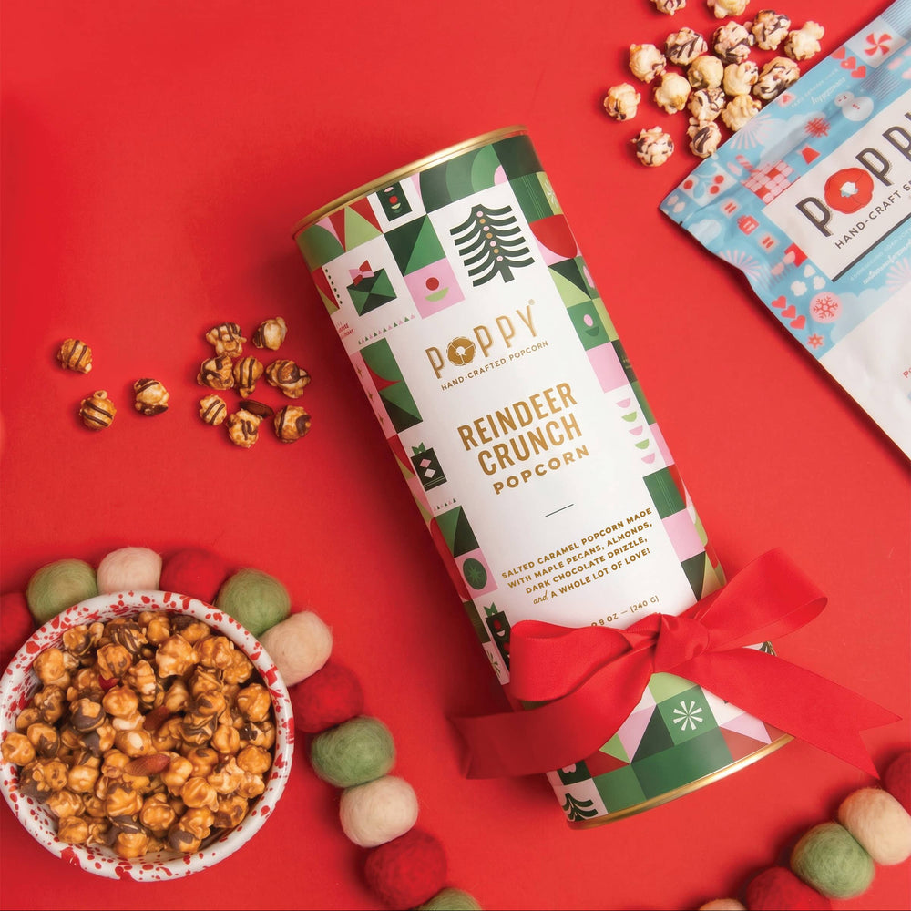 A cylindrical tin of Poppy Reindeer Crunch Popcorn sits on a bright red background, styled with scattered popcorn kernels, a bowl of caramel popcorn, and a blue bag of Poppy popcorn. The tin features a festive green, white, and pink holiday design with a red ribbon tied around the base.