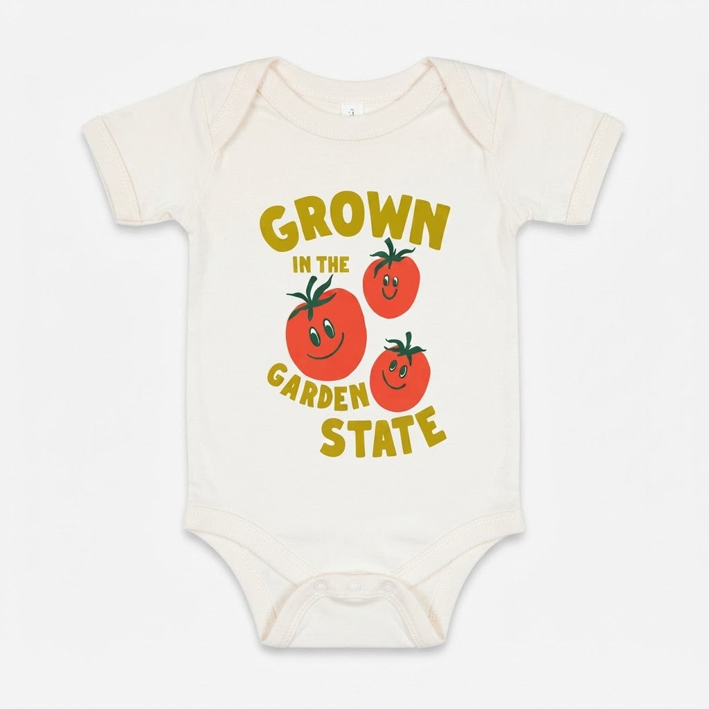 An off-white short-sleeve baby bodysuit featuring a colorful graphic print that reads 'GROWN IN THE GARDEN STATE' in yellow text around three smiling red tomatoes with green leaves.