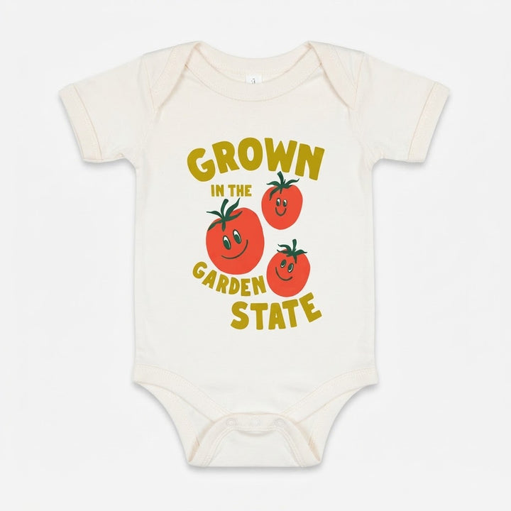 An off-white short-sleeve baby bodysuit featuring a colorful graphic print that reads 'GROWN IN THE GARDEN STATE' in yellow text around three smiling red tomatoes with green leaves.