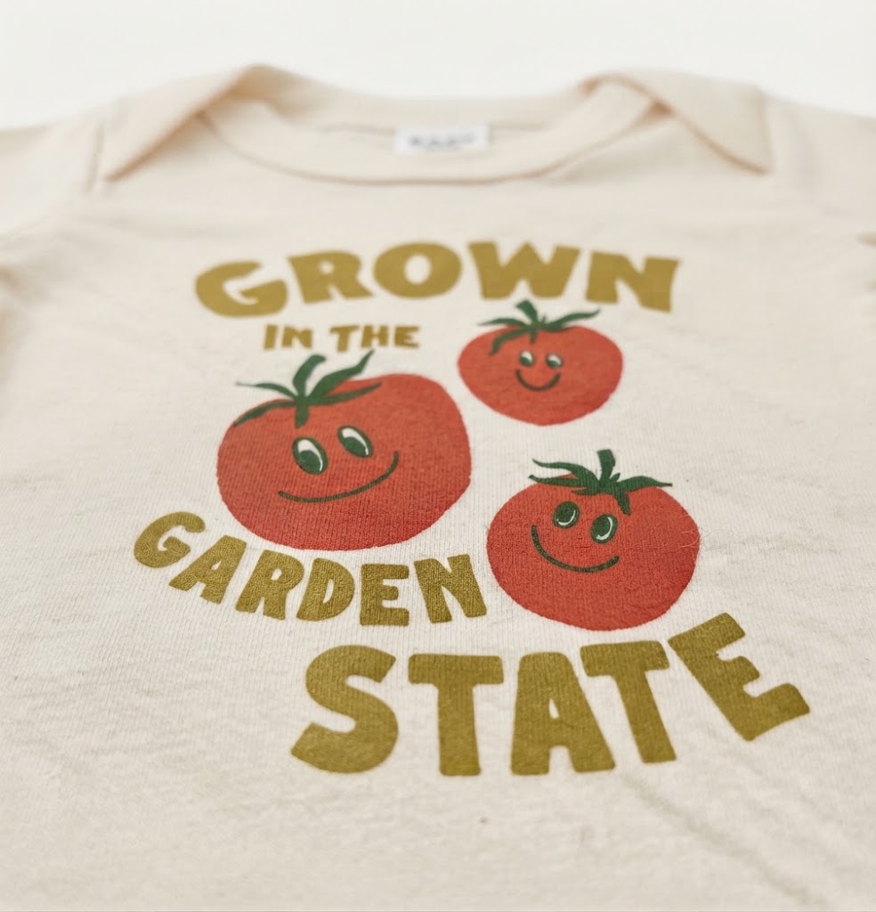 An off-white baby bodysuit featuring a colorful graphic print that reads 'GROWN IN THE GARDEN STATE' in gold lettering, accompanied by three smiling red tomatoes with green stems.