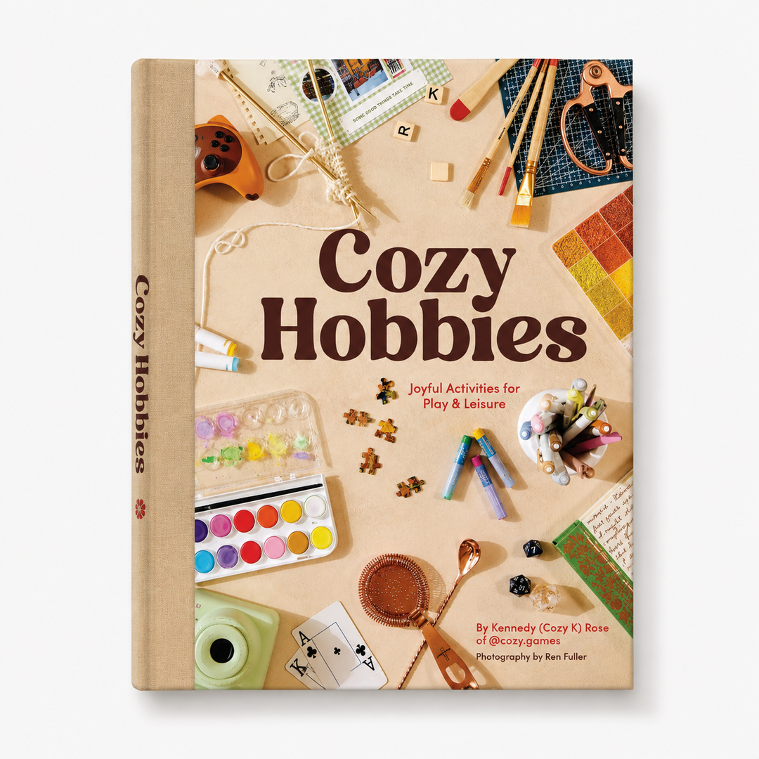 The book 'Cozy Hobbies' by Kenndee (Cozy K) Rose is shown standing upright. The cover features a warm beige background decorated with various colorful hobby items like paintbrushes, a watercolor palette, a mini camera, and cards.