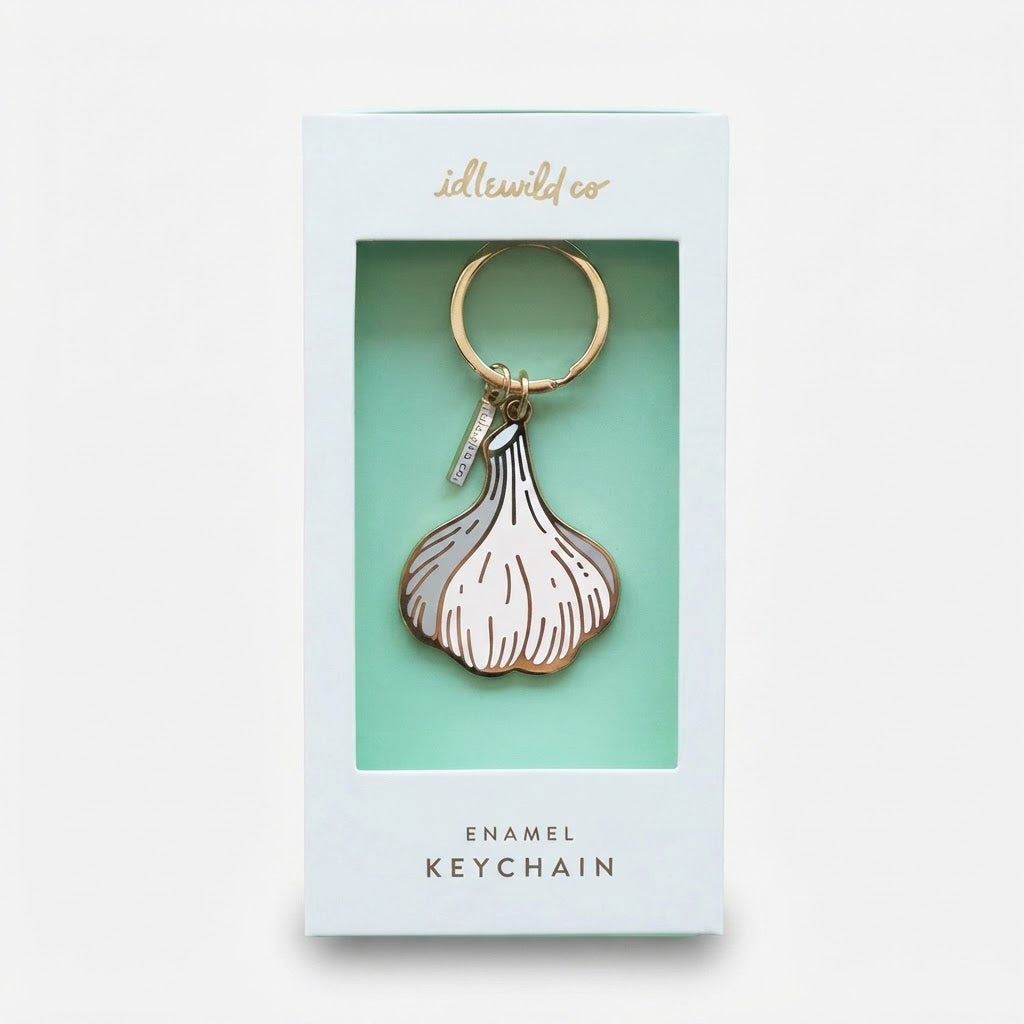 An enamel keychain shaped like a garlic bulb, presented inside its light blue and mint green retail packaging box. The keychain features a white enamel body with gold metal outlines and a gold split ring.