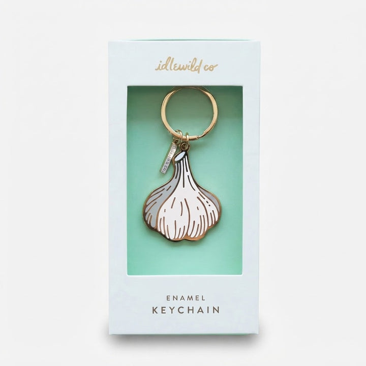 An enamel keychain shaped like a garlic bulb, presented inside its light blue and mint green retail packaging box. The keychain features a white enamel body with gold metal outlines and a gold split ring.