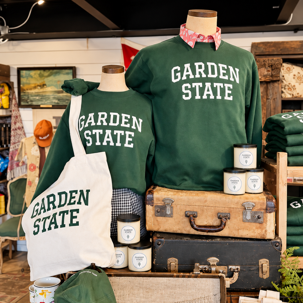 A retail display featuring two dark green crewneck sweatshirts and a matching tote bag printed with 'GARDEN STATE' in white, styled on mannequins and displayed with vintage suitcases, mugs, and small merchandise in a shop interior.