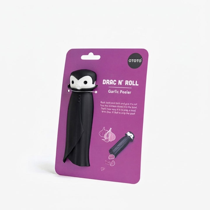 A garlic peeler shaped like a cartoon character in a black robe, mounted on a purple card backing. The card has white text reading 'OTOTO DRAC 'N' ROLL Garlic Peeler' and includes small garlic illustrations.