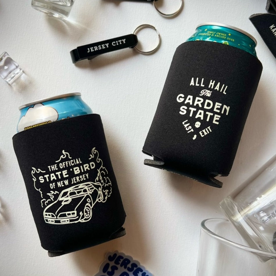 A photo of two cans wrapped in black koozies: One reads "The Official State Bird of New Jersey" with a picture of a car and flames  and the second koozie reads "All Hail the Garden State. Last exit" There is also a black Jersey City bottle opener in the photo.