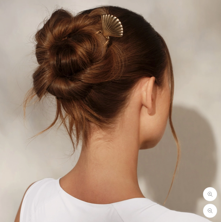 A woman shown from the back wearing a brown hair bun styled with a gold fan-shaped hair clip, against a neutral light gray background.