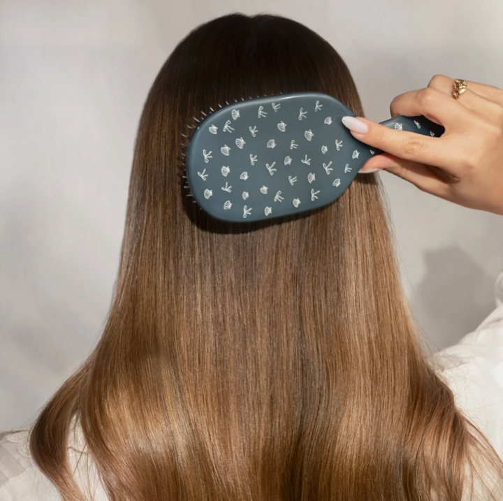A person with long, straight brown hair is shown from behind while being brushed with a dark blue-gray paddle hairbrush that has a white pattern of small icons on the bristle cover.
