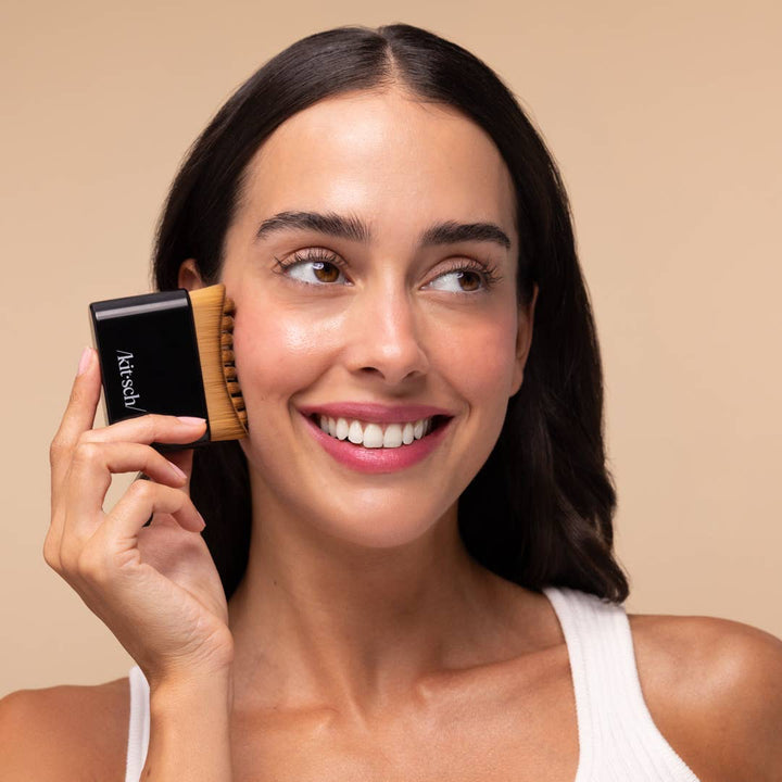 A woman with dark hair wearing a white ribbed tank top holds a black and wood facial cleansing brush against her cheek, set against a solid beige background.