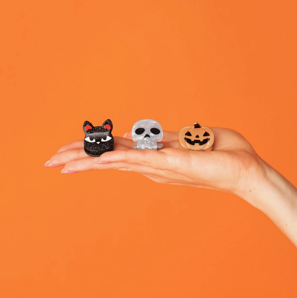 Three small Halloween-themed rings are displayed on a person's open hand against a solid orange background. The rings feature a black cat, a silver skull, and a jack-o'-lantern pumpkin design.