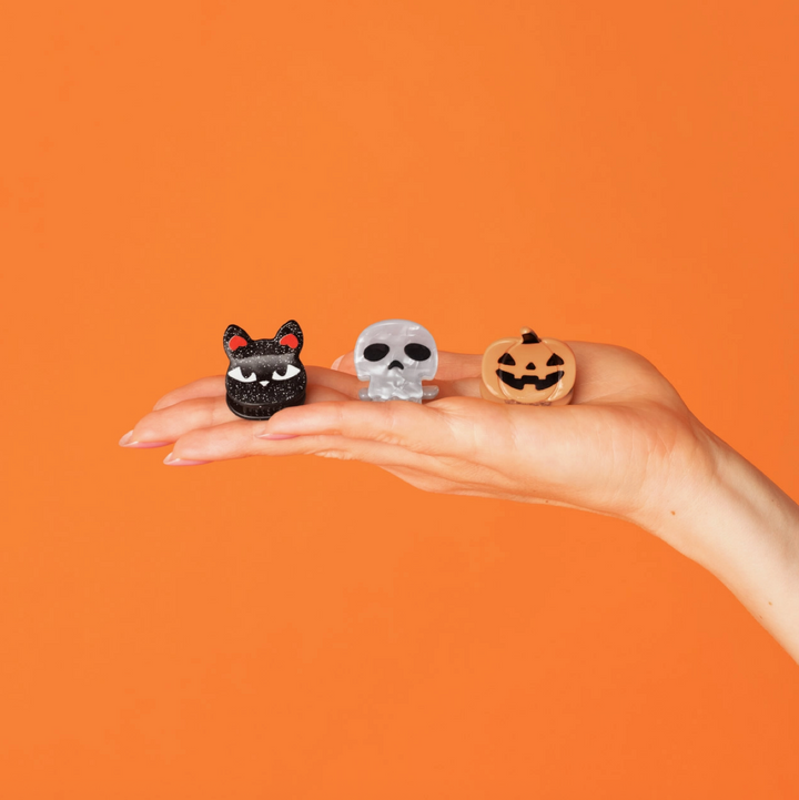 Three small Halloween-themed rings are displayed on a person's open hand against a solid orange background. The rings feature a black cat, a silver skull, and a jack-o'-lantern pumpkin design.