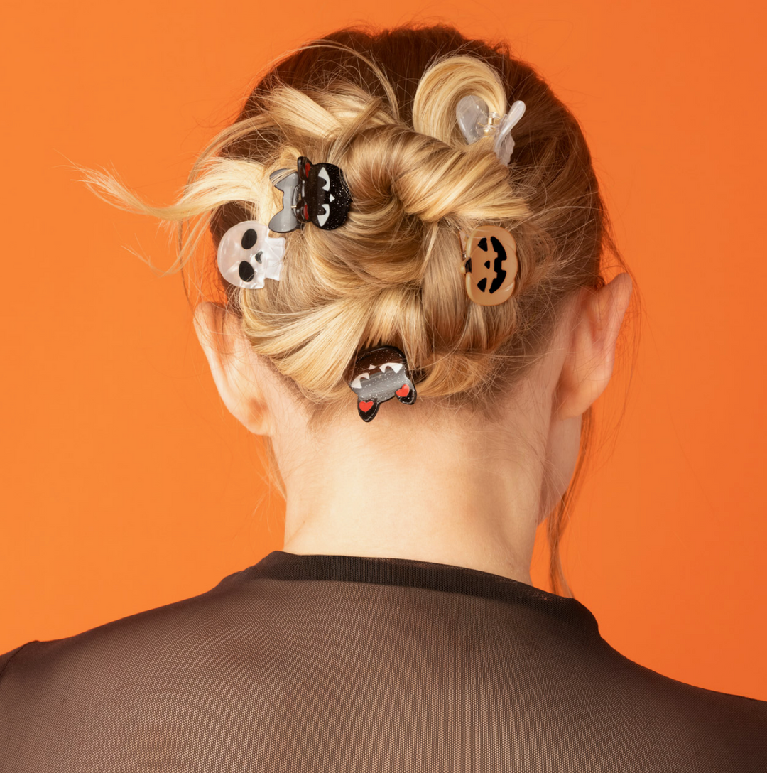 A person shown from the back with blonde hair styled in a messy bun, wearing six Halloween-themed hair clips against a solid orange background.