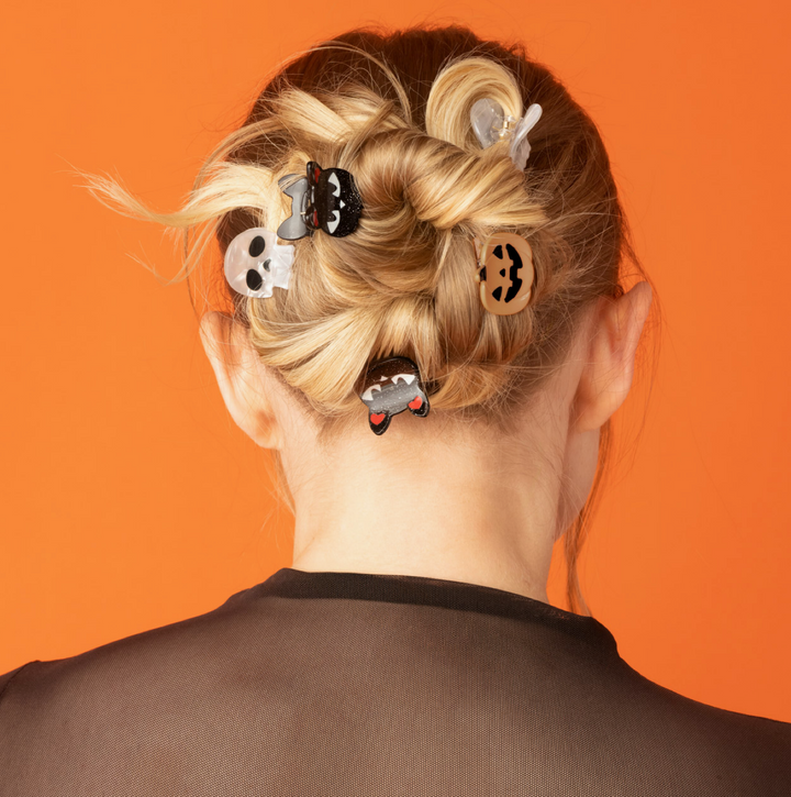 A person shown from the back with blonde hair styled in a messy bun, wearing six Halloween-themed hair clips against a solid orange background.