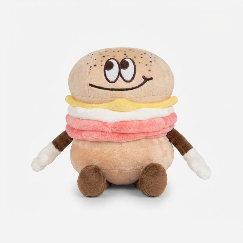 A plush toy shaped like a smiling hamburger with a sesame seed bun, yellow and pink cheese layers, white cream, and brown arms and legs, photographed on a light gray background.