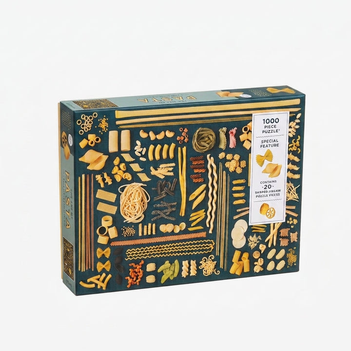 A 1000-piece jigsaw puzzle box featuring pasta illustrations in gold, yellow, and orange on a dark teal background, photographed on a light gray studio background.