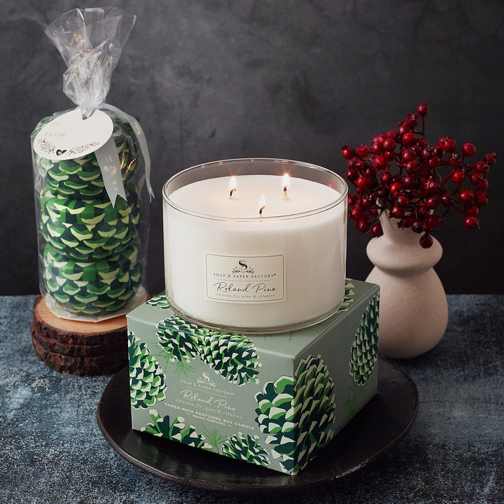 Candle with decorative packaging on a dark surface, surrounded by a pine cone package and red berries.