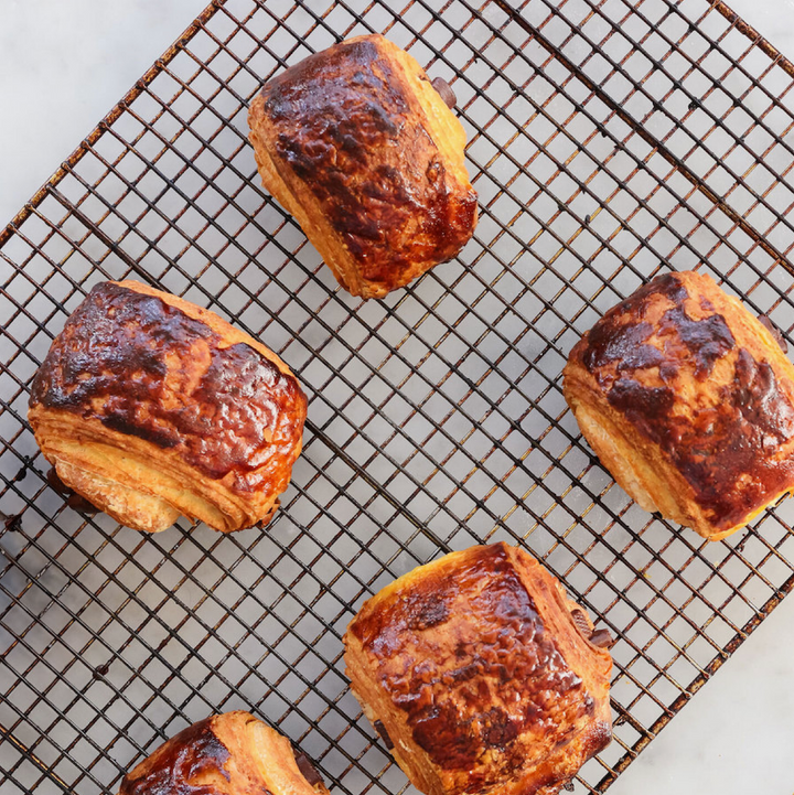 Five golden-brown flaky pastries with glossy caramelized tops arranged on a black wire cooling rack over a light marble surface.