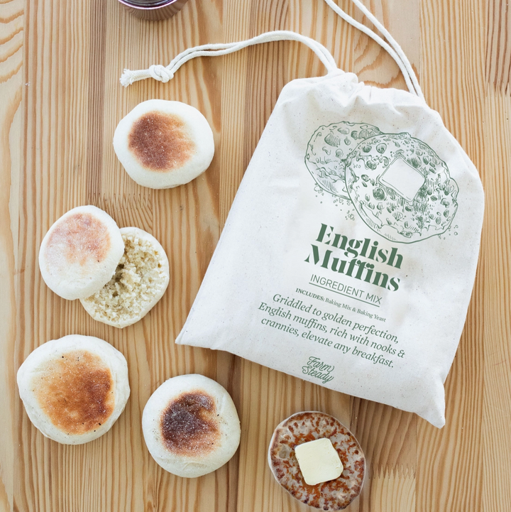 A white canvas drawstring bag labeled 'English Muffins INGREDIENT MIX' sits on a light wood tabletop, surrounded by several cooked English muffins, a small jar of dark red jam, and a muffin topped with butter. No person or animal is present, so this is Product/Packaging rather than Lifestyle.