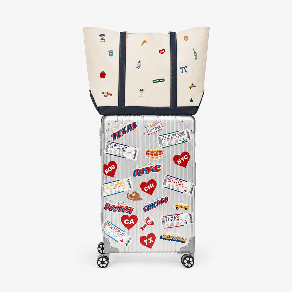 A cream canvas tote bag with navy straps and small multicolor travel-themed stickers, shown resting on top of a silver hard-shell suitcase covered in colorful destination-style stickers, against a light gray background.