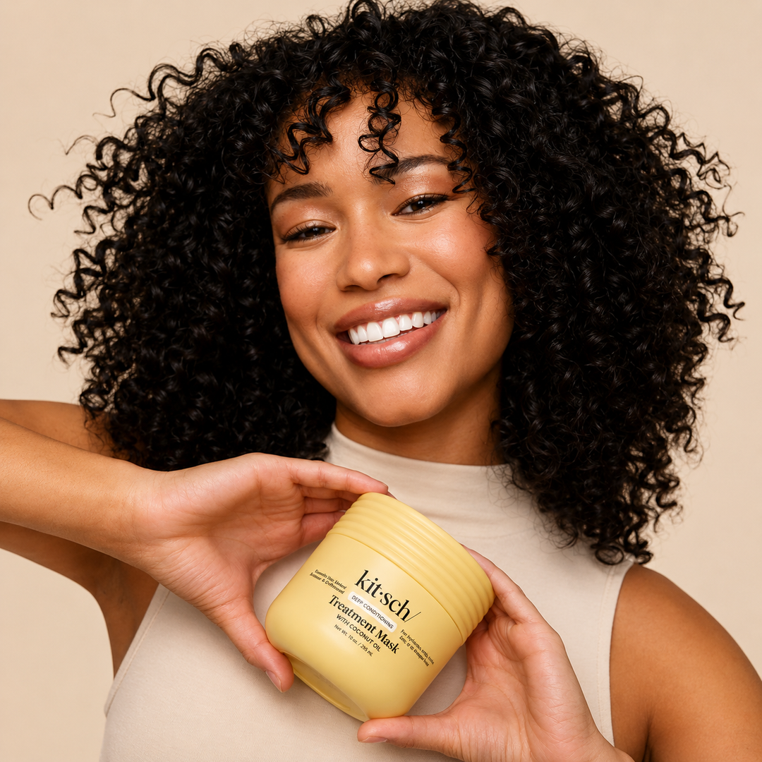A smiling woman with voluminous curly black hair holds a yellow jar of Kitsch Hair Treatment Mask against a warm beige background.