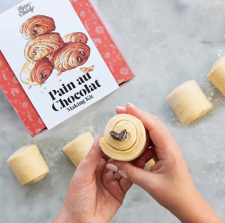 A hands-on view of a 'Pain au Chocolat Making Kit' by Fancy Beauty, showing a person's hands holding a rolled pastry with chocolate chips while the red and white kit box sits in the background on a light gray surface with other dough rolls scattered around.