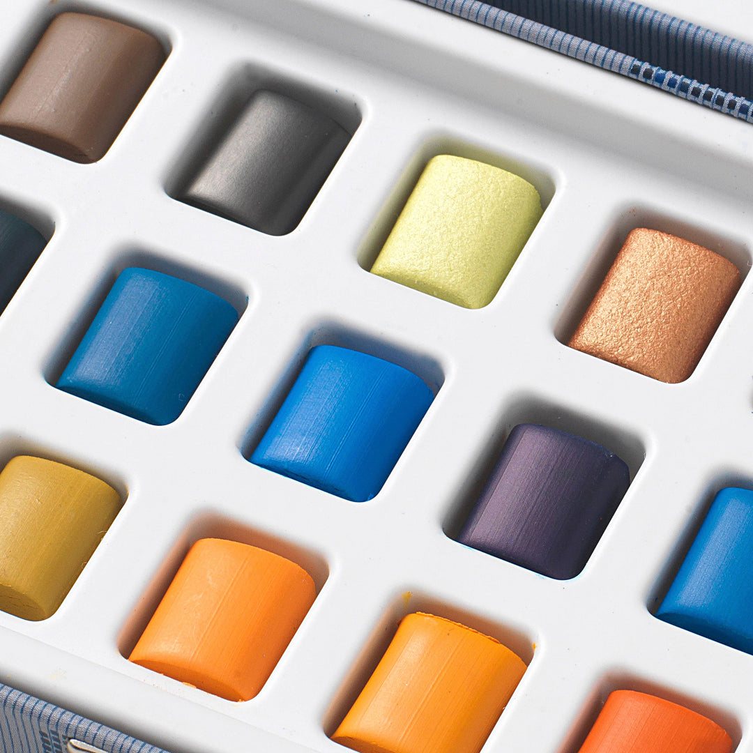 A close-up of a watercolor pastel set in a white plastic tray, showing multiple pastel sticks in assorted colors including blue, orange, yellow, purple, and brown.