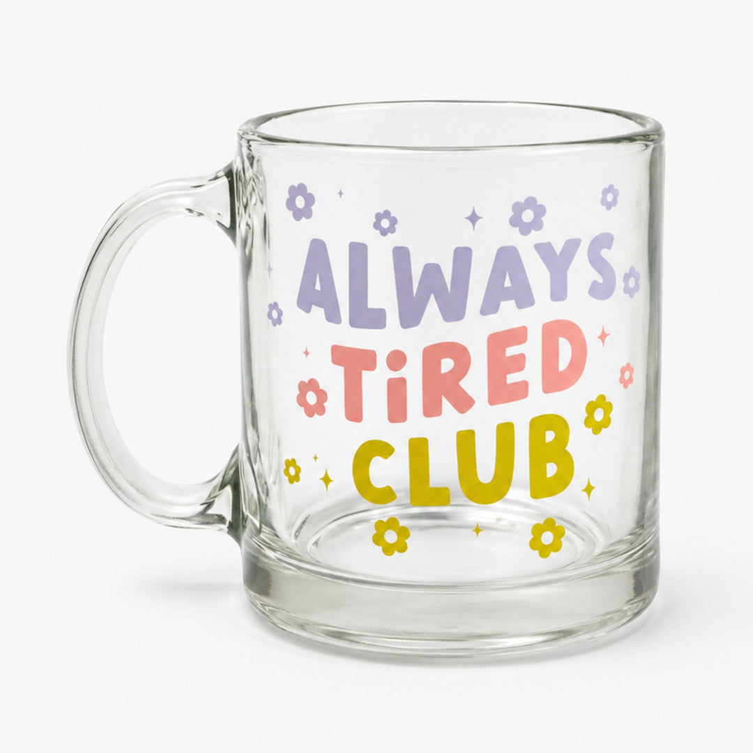A clear glass mug with a handle, featuring the text 'ALWAYS TIRED CLUB' in purple, pink, and yellow lettering, decorated with small colorful flowers and stars on a light gray background.