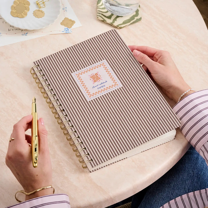 B5 hardcover spiral notebook with striped design held by a person with a pen, on a light table