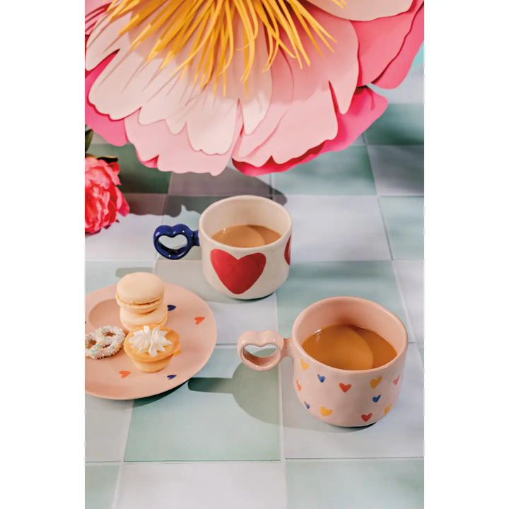 Danica big love coffee mug with heart-shaped handle beside colorful macarons and a floral background