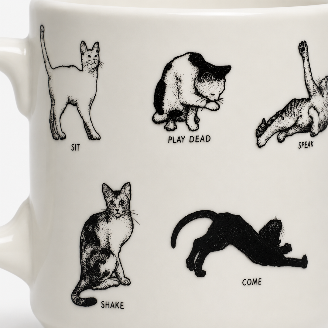 White ceramic mug with black line-art illustrations of cats in different poses and labels: SIT, PLAY DEAD, SPEAK, SHAKE, and COME.