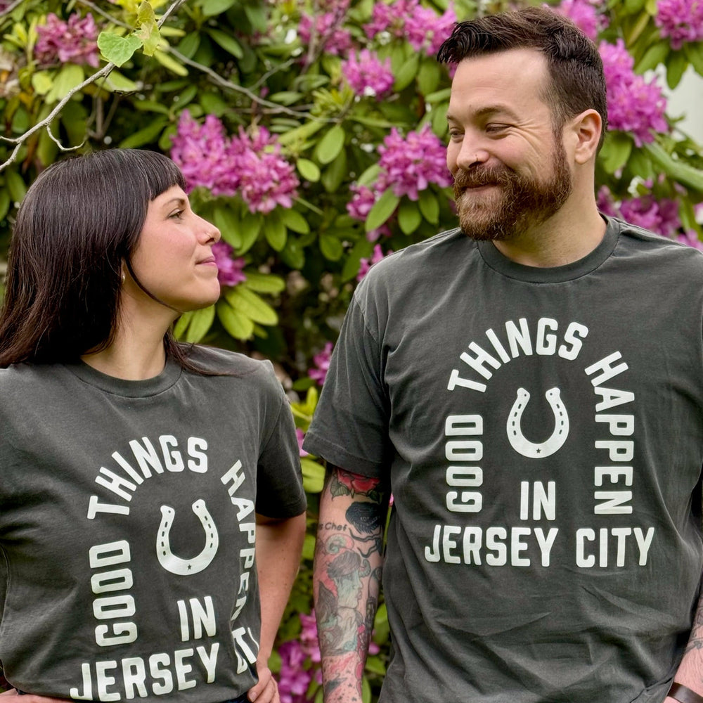 Man and woman wearing Good Things Happen in Jersey City shirt standing outdoors with purple flowers