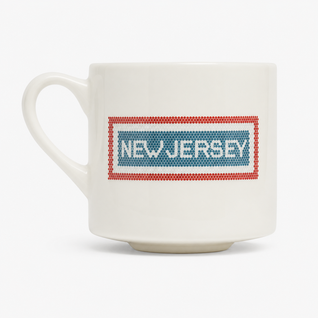 A white ceramic coffee mug featuring a printed graphic of the New Jersey state flag inside a red border, with 'NEW JERSEY' written in white text across the blue field.
