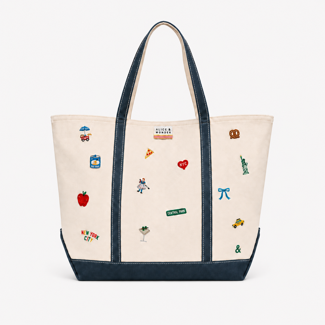 An off-white canvas tote bag with dark navy blue trim, handles, and bottom panel, decorated with various colorful embroidered New York City-themed motifs including a red apple, pizza slice, Statue of Liberty, and street signs.