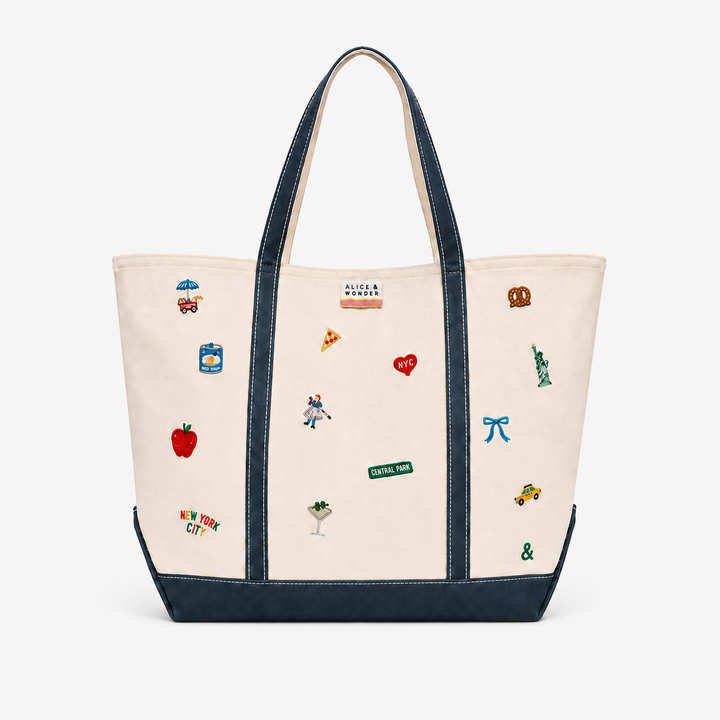 An off-white canvas tote bag with dark navy blue trim, handles, and bottom panel, decorated with various colorful embroidered New York City-themed motifs including a red apple, pizza slice, Statue of Liberty, and street signs.
