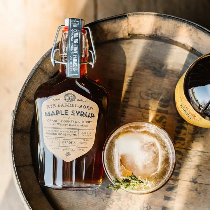 Finding Home Farms Rye Barrel Aged Maple Syrup in a glass bottle on a wooden barrel with a cocktail