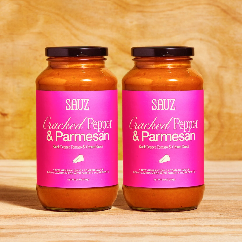 Two Sauz glass jars of cracked pepper parmesan pasta sauce with bright pink labels on a wooden surface