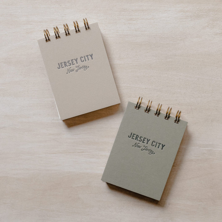 Two mini jotter notebooks: one tan and the other gray. Both read "Jersey City, New Jersey"