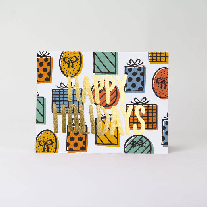 Gold foil HAPPY HOLIDAYS greeting card with illustrated presents in the background. Card is in a landscape layout