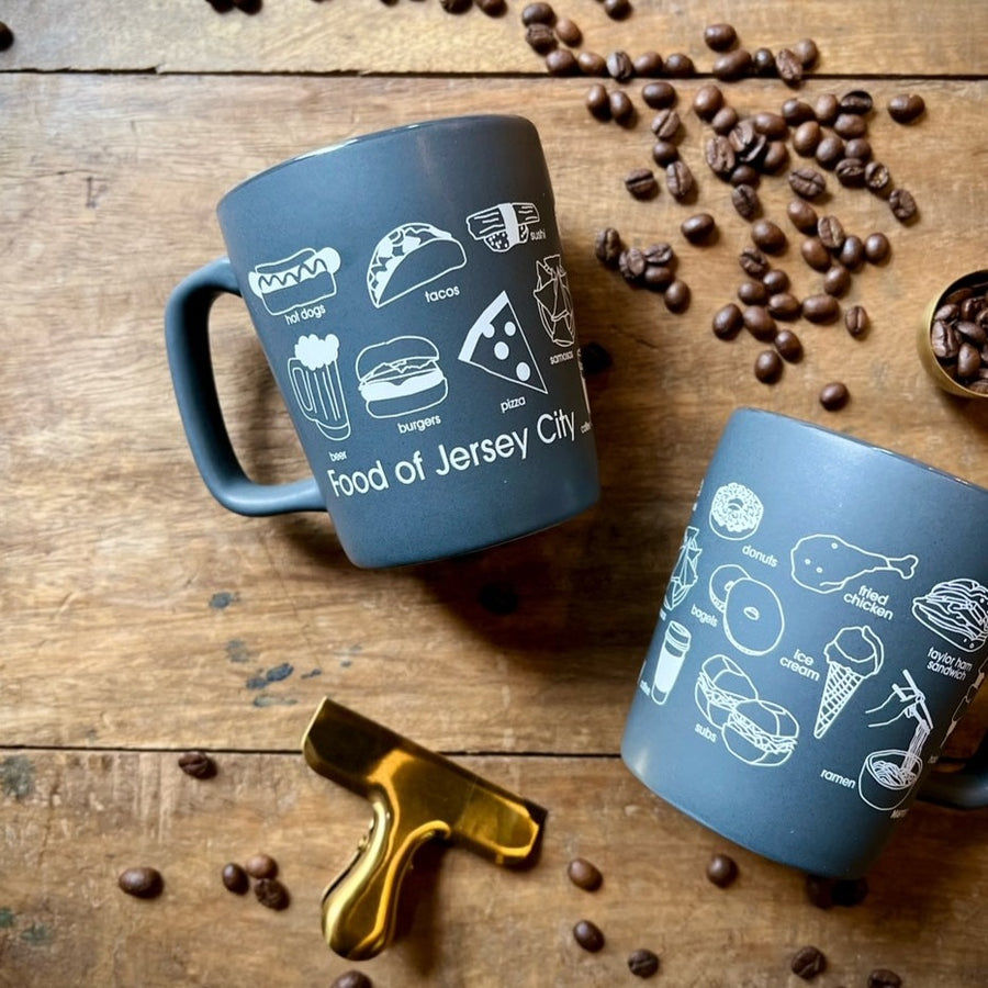 Two gray mugs surrounded by spilled coffee beans. The mugs read "Food of Jersey city" in white letters with outlines of various food items. Some of the food items include: Pizza, bagels, ice cream, burgers & beer.