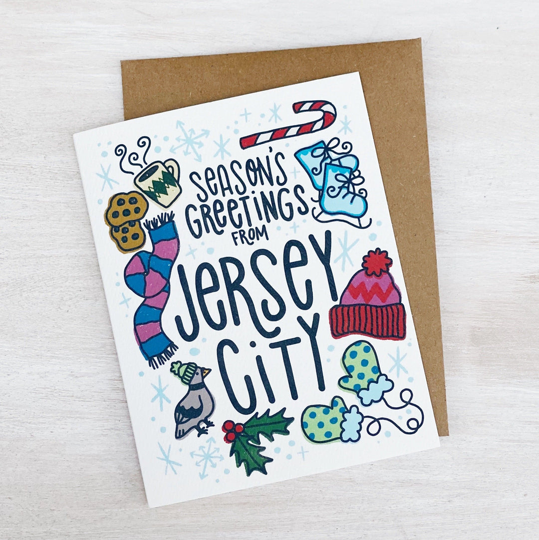 Rectangular card that reads "Season's Greetings from Jersey City" with a snowflake boarder Within the boarder there are illustrations of various winter themed things such as: a candy cane, ice skates, hat, gloves, scarf, a pigeon wearing a hat and finally a coffee with cookies.