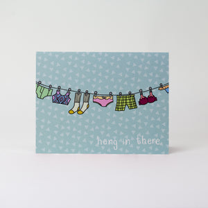Horizontal facing card with a sky blue background that has a clothesline with undergarments hanging from it. The card reads "hang in there"