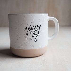 Two toned ceramic mug that reads "Jersey City" across the front. The bottom half of the mug is a tan color & the rest is white with black specks. 