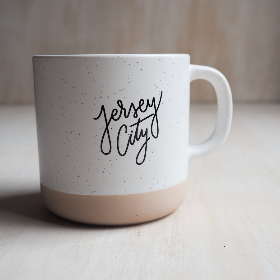 Two toned ceramic mug that reads "Jersey City" across the front. The bottom half of the mug is a tan color & the rest is white with black specks. 