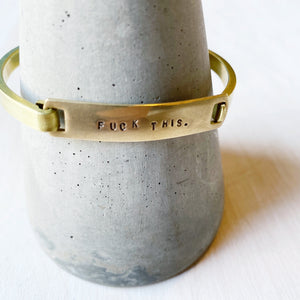 Brass cuff bracelet on grey cone.  Text reads "Fuck This."