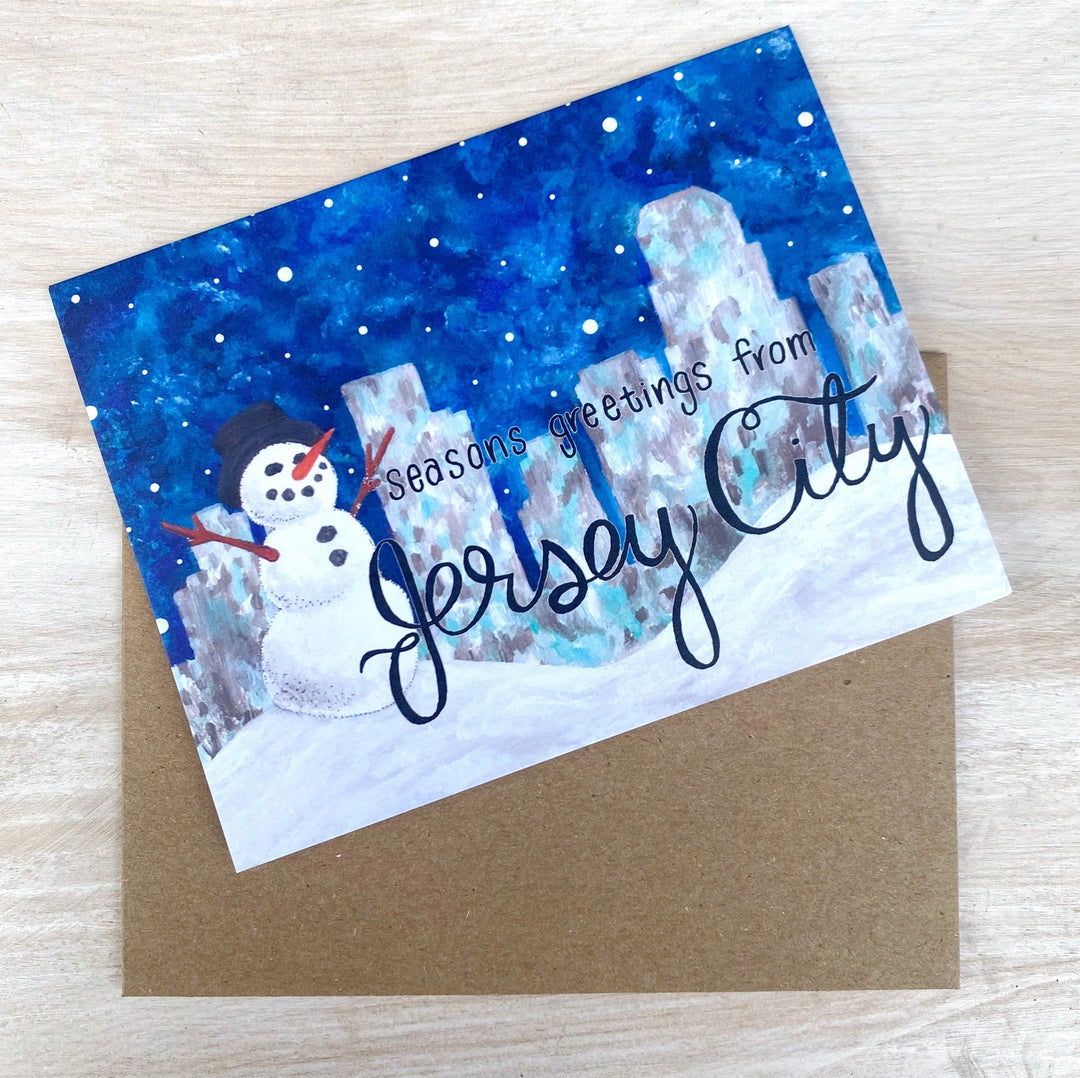 Rectangular card with a snowy night background that reads "Seasons greetings from Jersey City" The snowy night background includes a snowman wearing a top hat & a city skyline behind him.