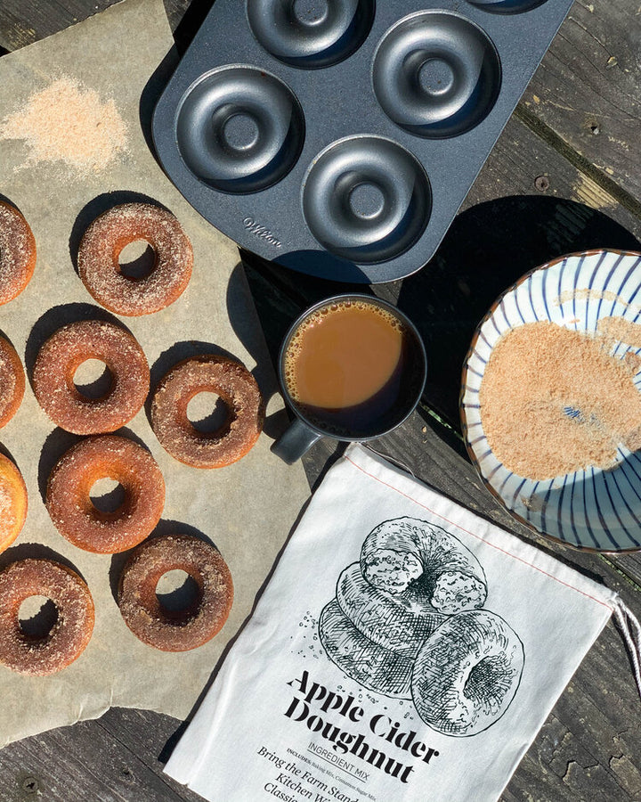 Apple cider doughnut baking kit with baked doughnuts, cinnamon sugar, and coffee on a rustic table