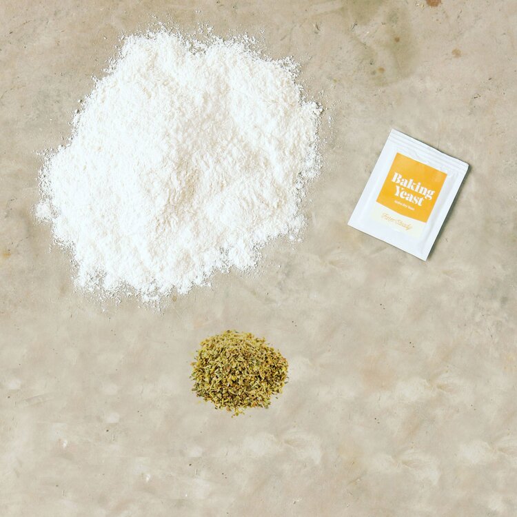 Garlic herb focaccia baking kit ingredients including flour, baking yeast, and dried herbs arranged on a marble surface