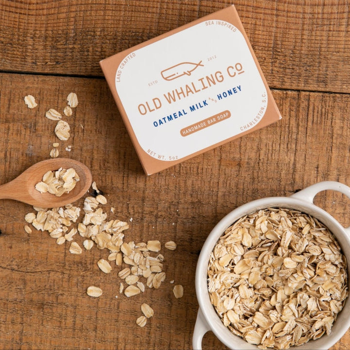 Old Whaling Co oatmeal milk honey bar soap next to a bowl and spoon of oats on wooden surface